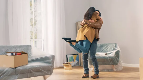 Pareja joven feliz y emocionada Mira a tu alrededor maravillado en su apartamento recién comprado / alquilado. Chica salta a los brazos de su novio abrazo. Gran hogar moderno brillante con cajas de cartón listo para desempacar . — Foto de Stock