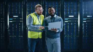 Sakallı siyah It mühendisi bir raf üzerinde çalışan sunucu bilgisayarları ile çalışan bir veri merkezi sunucu odası ortasında ayakta ve gülümsüyor.