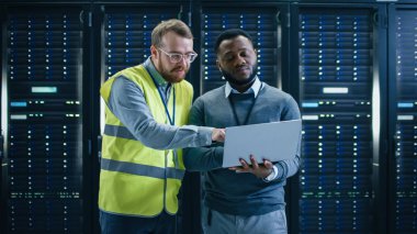 Sakallı o uzman gözlük ve yüksek görünürlük yelek ve siyah teknisyen meslektaşım bir laptop bilgisayar ile veri merkezi konuşan sunucu rafları yanında ayakta iken. Tanılama veya bakım çalıştırma.