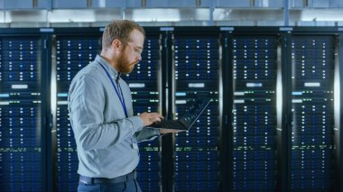 Sakallı veri merkezi o mühendis gözlük bir dizüstü bilgisayar ile sunucu raf koridoru üzerinden yürüyor. O çalışma sunucu dolapları görsel teftiş olduğunu.