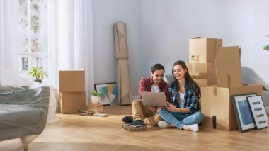 Yeni kira/satın daire kullanım dizüstü bilgisayar katında oturan mutlu genç çift. Ambalajsız karton kutular ve modern parlak Sweet Home kapalı mobilya.