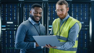 Sakallı o uzman gözlük ve yüksek görünürlük yelek ile bir laptop bilgisayar ve siyah teknisyen meslektaşım konuşan veri merkezi sunucu rafları yanında ayakta iken. Tanılama veya bakım çalıştırma.