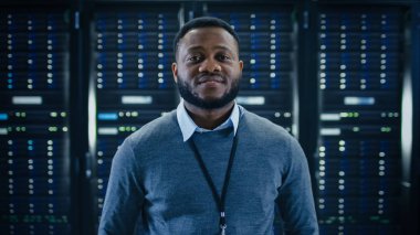 Sakallı siyah mühendis ayakta ve fotoğraf makinesinde çalışan bir veri merkezi sunucu odasının ortasında bir raf üzerinde çalışan sunucu bilgisayar.