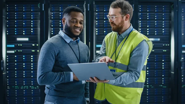 Bearded IT Specialist in Glasses and High Visibility Vest with a Laptop Computer and Black Technician Colleague Talking in Data Center while Standing Next to Server Racks. Running Diagnostics or Maintenance.