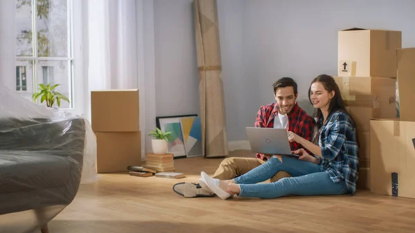 Joyeux jeune couple assis sur le plancher de l'appartement nouvellement loué / acheté Utilisez ordinateur portable. Boîtes en carton déballées et meubles recouverts dans la maison douce lumineuse moderne . — Photo