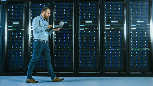 Bearded Data Center IT Professional Walking Through Server Rack Corridor with a Laptop Computer. He Stops and Wirelessly Inspects Working Server Cabinets.