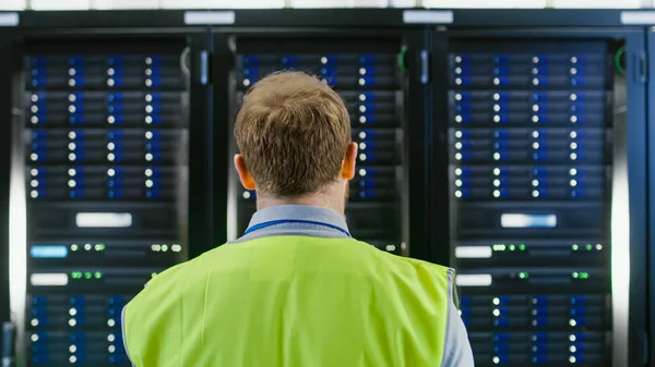 Back Shot of an IT Administrator in High Visibility Vest Walking Towards a Server Rack in Data Center Room.