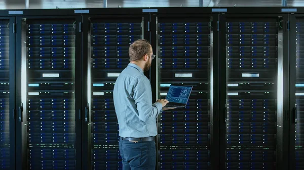 Der bärtige Brillenspezialist arbeitet am Laptop im Rechenzentrum, während er vor dem Servergestell steht. Durchführung von Diagnosen oder Wartungsarbeiten. — Stockfoto