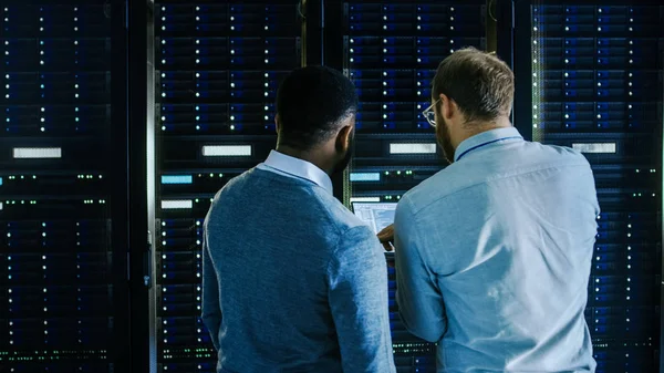 Brodaty IT technika w okularach z laptopem i czarny mężczyzna inżynier kolega rozmawiają w centrum danych podczas pracy obok szafy serwerowe. Uruchamianie diagnostyki lub wykonywanie prac konserwacyjnych. — Zdjęcie stockowe