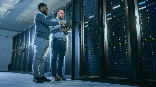 Bearded IT Technician in Glasses with a Laptop Computer and Black Male Engineer Colleague are Talking in Data Center while Working Next to Server Racks. Running Diagnostics or Doing Maintenance Work.