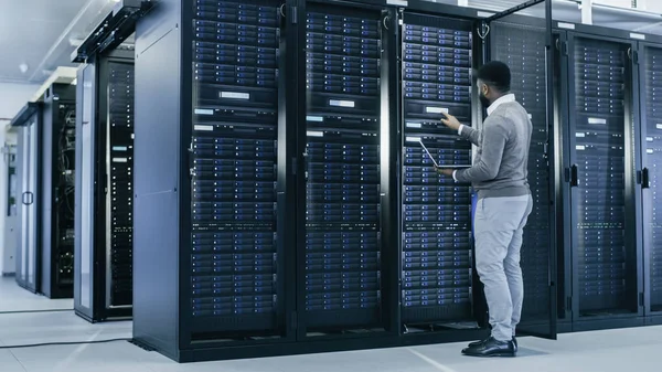 Especialista em TI preto está trabalhando no laptop no data center enquanto está em pé antes da cremalheira do servidor. Executando diagnósticos, colocando em dados ou fazendo o trabalho de manutenção . — Fotografia de Stock