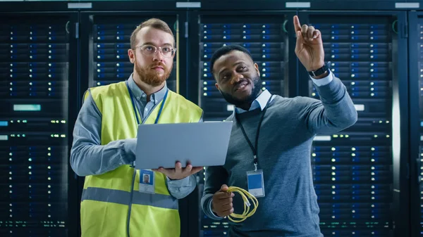 Especialista em TI em Óculos e Colete de Alta Visibilidade com Computador Laptop e Colega Técnica Preta Falando em Data Center enquanto está ao lado de Racks de Servidor. Executando diagnósticos e olhando para cima . — Fotografia de Stock