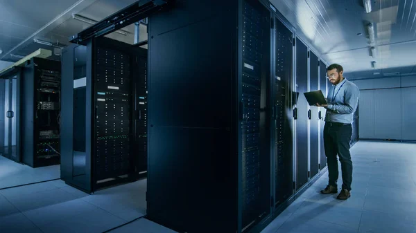 Bearded IT Specialist in Glasses is Working on Laptop in Data Center while Standing Before Server Rack. Running Diagnostics or Doing Maintenance Work. — Stock Photo, Image