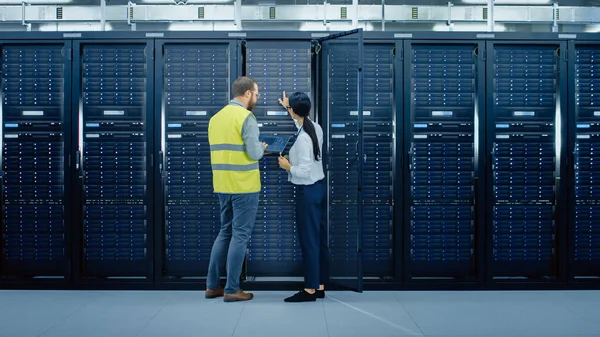 IT Admin in Glasses and High Visibility Vest with a Laptop Computer and Young Technician Colleague Talking in Data Center while Standing Next to Server Racks. Запуск диагностики или технического обслуживания . — стоковое фото