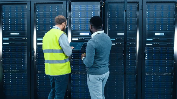 IT Administrator with a Laptop Computer in High Visibility Vest and Black Male Engineer are Talking in Data Center while Standing Before a Server Rack. Running Diagnostics or Doing Maintenance Work.