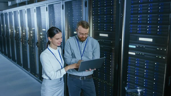 Skægget IT-tekniker i briller med en bærbar computer og smukke unge ingeniør kollega taler i datacenter, mens du arbejder ved siden af Server Racks. Kører diagnostik eller udfører vedligeholdelsesarbejde . - Stock-foto