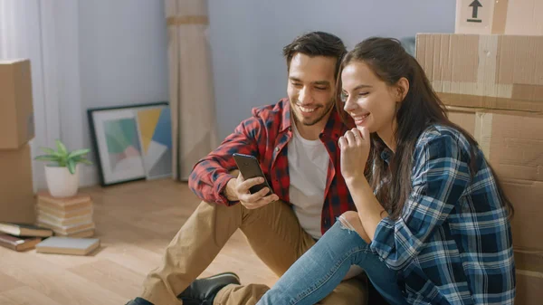 Boldog fiatal pár ül a padlón az újonnan bérelt/vásárolt apartman és használata smartphone. Csomagolatlan karton dobozok és fedett bútor a modern Bright Sweet Home. — Stock Fotó