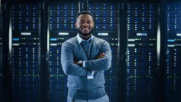 Engenheiro de TI preto barbudo em pé e posando com braços cruzados no meio de uma sala de servidor de data center de trabalho com computadores de servidor trabalhando em uma cremalheira . — Fotografia de Stock