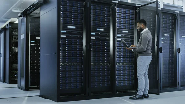 Especialista negro de TI está trabajando en el ordenador portátil en el centro de datos mientras está de pie antes del rack del servidor. Ejecución de diagnósticos, puesta en marcha de datos o trabajos de mantenimiento . —  Fotos de Stock