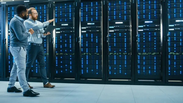 Técnico de TI con una computadora portátil y un compañero de ingeniero negro están hablando en el centro de datos mientras caminan junto a los bastidores del servidor. Ejecución de diagnósticos o trabajos de mantenimiento . —  Fotos de Stock