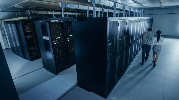 Acima do tiro largo de um administrador de TI com um computador portátil e um colega técnico novo que anda ao lado das cremalheiras do servidor no centro de dados. Executando diagnósticos ou manutenção . — Fotografia de Stock