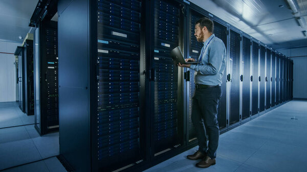 Bearded IT Specialist in Glasses is Working on Laptop in Data Center while Standing Before Server Rack. Running Diagnostics or Doing Maintenance Work.