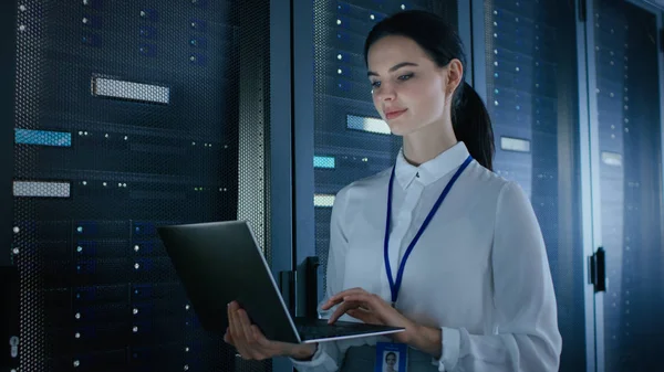 Ingeniera de TI femenina segura y hermosa está trabajando en el ordenador portátil en el centro de datos mientras está de pie ante el estante del servidor. Ejecución de diagnósticos o trabajos de mantenimiento . —  Fotos de Stock