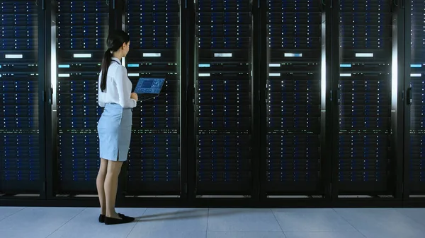 Bellissimo Data Center tecnico IT femminile che cammina attraverso il Corridoio dello scaffale del server con un computer portatile. Lei si ferma e visivamente Inspects lavoro armadi server . — Foto Stock