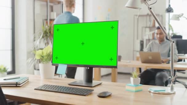 モダンクリエイティブオフィスの机の上に立つモックアップスクリーンデスクトップコンピュータ。若い専門家の多様なチームがコンピュータ上で働く背景明るいスタジオアパートで — ストック動画