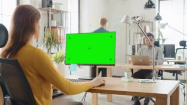 Omuz Over: Creative genç Woman, mock-up yeşil ekran ile masaüstü bilgisayar kullanarak onun masasında oturuyor. Arka plan parlak ofiste genç profesyoneller çeşitli takım bilgisayarlarda çalışma