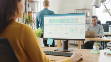 Omuz Over: yaratıcı genç kadın masası, proje yönetimi, Istatistik ve grafikler gösteren ekran ile masaüstü bilgisayar kullanarak oturan. Çalışma uzmanları çeşitli ekibi ile ofis