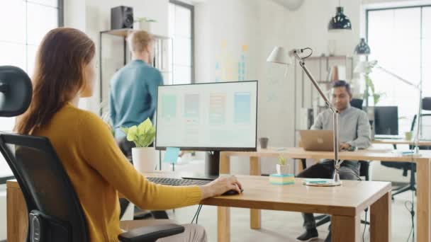 어깨 너머: 여성 모바일 소프트웨어 개발자는 스마트 폰 응용 프로그램 UI / Ux. 작업 전문가의 다양한 팀과 사무실을 보여주는 화면과 데스크톱 컴퓨터를 사용하여 그녀의 책상에 앉아 — 비디오