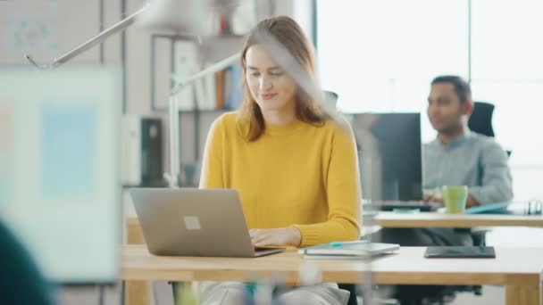 Schöne und kreative junge Frau, die an ihrem Schreibtisch sitzt und Laptop benutzt. im Hintergrund helles Büro, in dem ein vielfältiges Team junger Fachkräfte arbeitet — Stockvideo