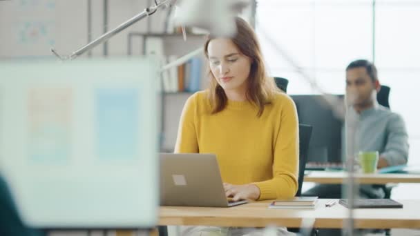 Krásná a kreativní mladá žena sedí u svého stolu pomocí přenosného počítače. Na pozadí světlý úřad, kde pracuje rozmanitý tým mladých profesionálů — Stock video