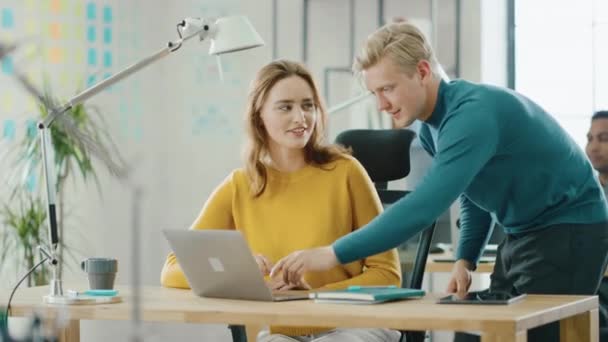 Mujer joven hermosa y creativa sentada en su escritorio usando una computadora portátil, Young Handsome Team Leader da consejos sobre los detalles del proyecto. Oficina donde trabajan diversos equipos de jóvenes profesionales — Vídeo de stock