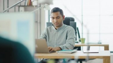 Akıllı ve yakışıklı Hint bilgi teknolojisi uzmanı masasında oturan bir laptop çalışır. Arka planda modern ofis genç profesyoneller çeşitli takım çalışma