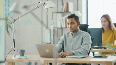 Akıllı ve yakışıklı Hint bilgi teknolojisi uzmanı masasında oturan bir laptop çalışır. Arka planda modern ofis genç profesyoneller çeşitli takım çalışma