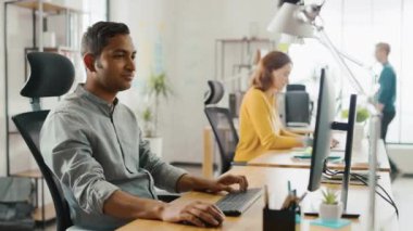 Akıllı ve yakışıklı Hintli ofis Işçisi onun masasında oturan bir laptop çalışır. Arka planda modern ofis genç profesyoneller çeşitli takım çalışma