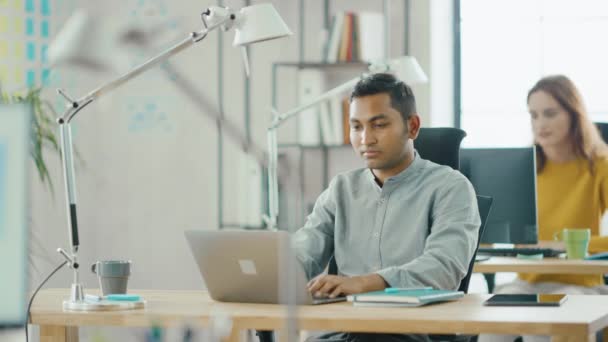 Розумний і красивий індійський фахівець з інформаційних технологій сидить на своєму столі працює на ноутбуці. У фоновому режимі сучасний офіс з різноманітною командою молодих професіоналів, що працюють — стокове відео