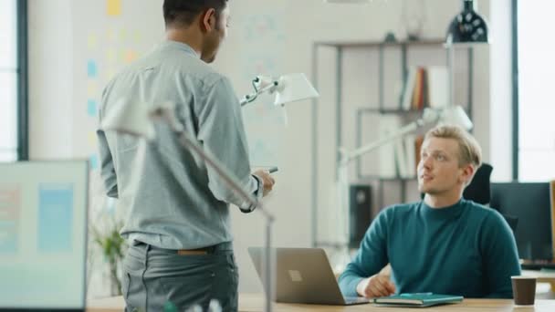 El apuesto especialista de TI sentado en su escritorio trabaja en una computadora portátil, habla con el gerente de proyectos profesional. Equipo Diverso de Jóvenes Profesionales que Trabajan en la Oficina — Vídeo de stock