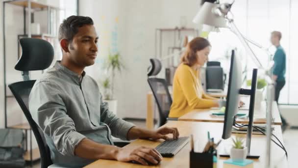 Smart och vacker indisk kontorsarbetare sitter vid sitt skrivbord fungerar på en bärbar dator. I bakgrunden moderna kontor med olika team av unga yrkesverksamma som arbetar — Stockvideo