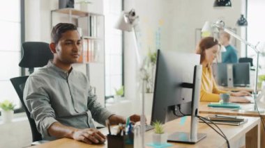 Akıllı ve yakışıklı Hintli ofis Işçisi onun masasında oturan bir laptop çalışır. Arka planda modern ofis genç profesyoneller çeşitli takım çalışma