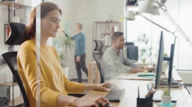 Güzel ve yaratıcı genç kadın oturma onu masa kullanarak laptop bilgisayar. Arka plan parlak ofiste genç profesyoneller çeşitli takım çalışma