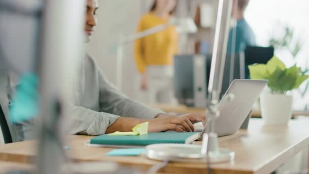 Trabajador de oficina indio inteligente y guapo sentado en su escritorio trabaja en una computadora portátil. En segundo plano Oficina moderna con equipo diverso de jóvenes profesionales que trabajan — Vídeos de Stock