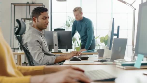 Beau spécialiste de la TI assis à son bureau travaille sur un ordinateur portable, a une discussion avec le gestionnaire de projet indien. Une équipe diversifiée de jeunes professionnels qui travaillent au bureau — Video