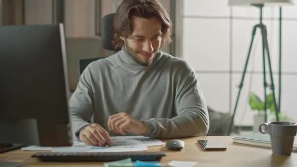 ハンサムな長い髪の起業家デスクトップコンピュータのオフィスの仕事で彼の机に座って、文書、グラフを扱う。スマートフォン、ソーシャルメディアアプリ、メールの書き込み、メッセージの使用 — ストック動画