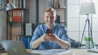 Çalışma masasında oturan yaratıcı girişimci, şık ofisteki bir dizüstü bilgisayarda çalışıyor, Sosyal Medya Uygulamaları için akıllı telefon kullanıyor, e-posta şirketleri, Haberler okuyor, İnternet 'e göz atıyor