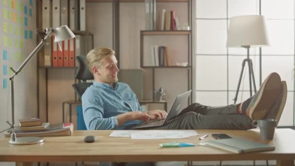 Knappe jonge ondernemer werkt op een laptop met zijn benen op het bureau. Ontwikkelaar Gebruikt Notebook om Software Unicorn Startup te maken. Chill Student Writing Paper voor de universiteit — Stockvideo