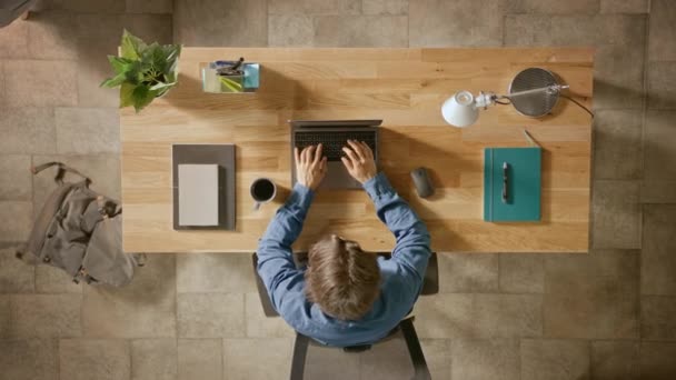 Zoom in der Aufnahme von oben: Geschäftsmann sitzt am Holztisch und arbeitet an einem Laptop in seinem Homeoffice. er trinkt Kaffee, tippt, schreibt E-Mails, surft im Internet, entwirft Software, kauft online ein — Stockvideo
