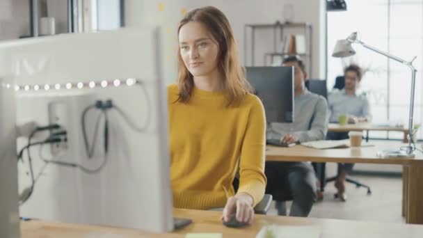 Bella e intelligente dai capelli rossi specialista femminile seduta alla sua scrivania lavora su un computer desktop. Sullo sfondo il moderno ufficio luminoso con diversi gruppi di professionisti che lavorano per la crescita delle startup — Video Stock